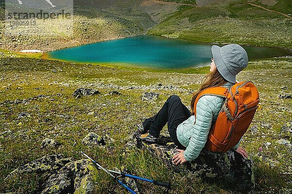 Nette Frau in einem Panama mit einem Rucksack sitzt auf einem Stein und ruht sich nach einem Trekking in den Bergen aus  in der Nähe des Sees