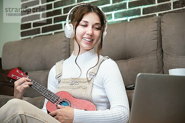 Fröhliches kaukasisches Mädchen  das Ukulele spielt und in einen Laptop schaut  sitzt auf dem Boden neben dem Sofa in der Wohnung. Junge Frau  die lernt  wie man akustische Ukulele während des Tages zu Hause spielt