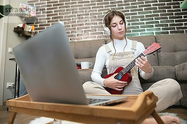 Schöne junge Hipster Mädchen in Freizeitkleidung lernen  die Ukulele Gitarre zu spielen  während auf dem Boden im Wohnzimmer zu Hause sitzen  Hobby Freizeitkonzept