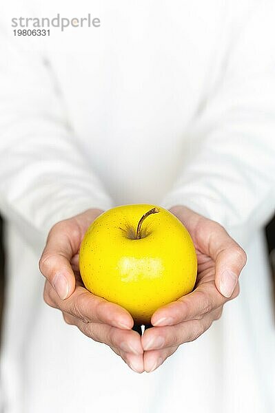 Die Hände einer jungen Frau halten einen gelben Apfel vor dem Hintergrund ihrer selbst in einem weißen Pullover. Frische Früchte. Pastellfarben. Nahaufnahme  Draufsicht