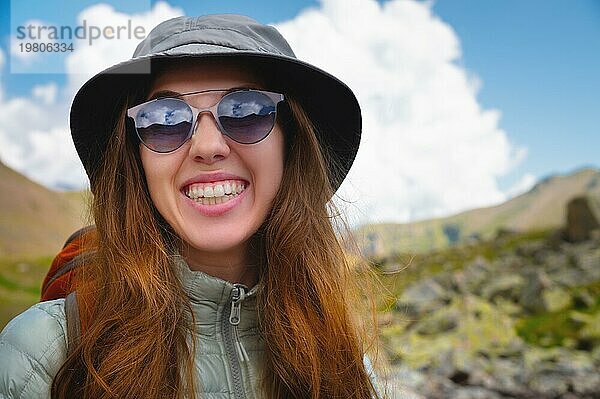 Lächelndes junges Mädchen vor einem blaün  bewölkten Himmel und Bergen. Porträt einer glücklichen Frau Tourist in Sonnenbrille und panama