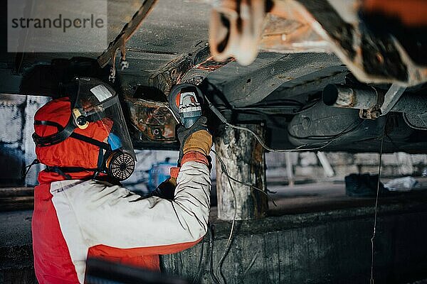 Männlicher Autowerkstattmitarbeiter reinigt den Boden der Karosserie mit einer Schleifmaschine. Imprägnieren von Metall einer Autokarosserie  Autoreparatur