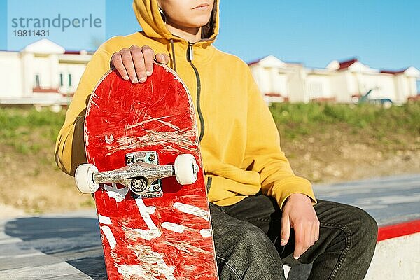 Nahaufnahme eines jungen Hipsters in Sweatshirt und Jeans  der traurig in einem Skatepark sitzt und ein Skateboard hält