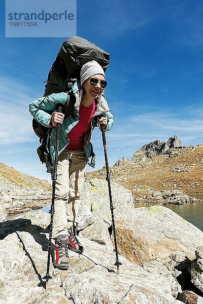 Schlankes und sympathisches Wandermädchen mit Sonnenbrille und blaür dünner Daunenjacke mit Rucksack und Trekkingstöcken steigt auf einen hohen Felsen vor dem Hintergrund von Felsen und einem hohen Bergsee