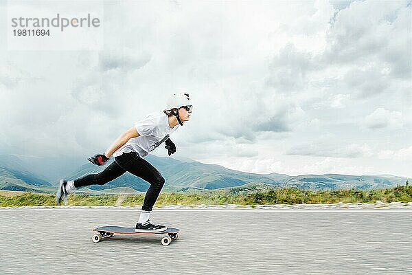 Ein junger Longboarder schiebt seinen Fuß auf seinem Longbord über die Landstraße vor dem Hintergrund der kaukasischen Landschaft