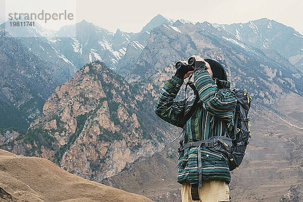 Ein Hipster Mann mit Bart  Hut  Jacke und Rucksack in den kaukasischen Bergen hält ein Fernglas in den Händen und schaut in die Ferne  Abenteuer  Tourismus  Tracking
