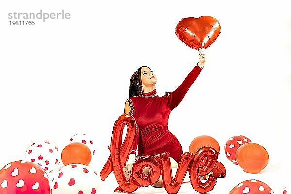 Schöne junge Frau in rotem Kleid umgeben von Luftballons auf weißem Hintergrund mit Liebe Zeichen. Valentinstag Konzept. Rote und weiße Farbpalette. Kopieren Raum