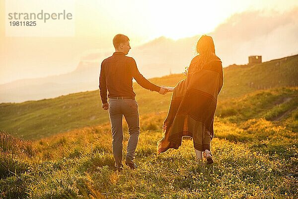 Ein junges Paar. Der Mann führt ein lockiges  in ein Plaid gehülltes Mädchen mit einem Blumenstrauß bei Sonnenuntergang. Das Konzept des Vertrauens und der glücklichen jungen Familie