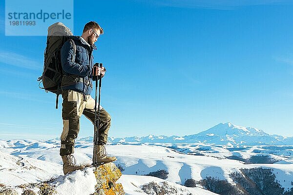 Ein Hipster Reisender mit Bart und Sonnenbrille in der Natur. Ein Mann wandern in den Bergen mit einem Rucksack und skandinavischen Wanderstöcke im Hintergrund eines Berges Elbrus Landschaft und blaün Himmel. Reisen Lifestyle Abenteuer Outdoor Erholung übertreffen