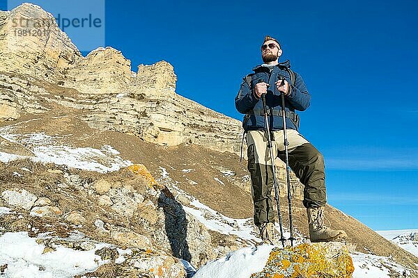 Ein Hipster Reisender mit Bart und Sonnenbrille in der Natur. Ein Mann wandert in den Bergen mit einem Rucksack und skandinavischen Wanderstöcken vor dem Hintergrund einer Berglandschaft und blauem Himmel. Reisen Lifestyle Abenteuer Outdoor Erholung Outdoor Sport