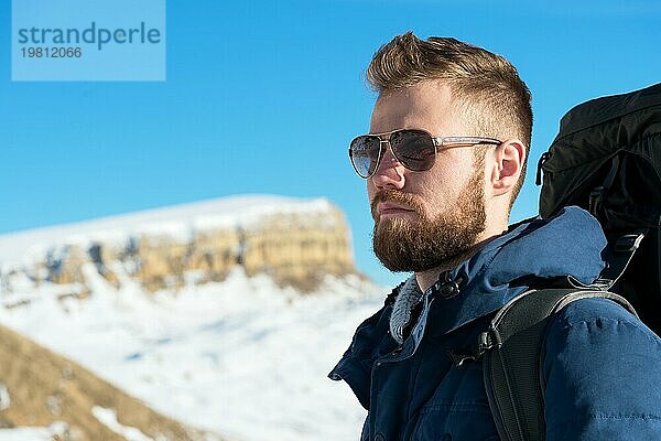 Ein Hipster Reisender mit Bart und Sonnenbrille in der Natur. Ein Mann wandert in den Bergen mit einem Rucksack und skandinavischen Wanderstöcken vor dem Hintergrund einer Berglandschaft und blauem Himmel. Reisen Lifestyle Abenteuer Outdoor Erholung Outdoor Sport