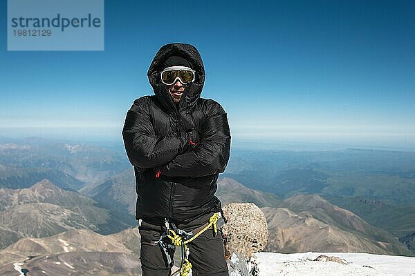 Bergsteiger auf dem Gipfel. Ein ausgerüsteter Bergsteiger mit Skimaske und Daunenjacke mit Kapuze  dem auf dem Gipfel kalt ist. Das Konzept der Entspannung in den Bergen