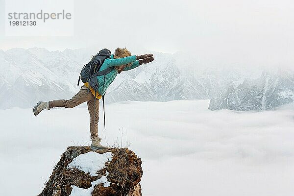 Ein fröhlich lächelndes Hipster Mädchen reist in einer Daunenjacke mit einem Rucksack und einem großen Pelzhut und steht in einer Superman Flugposition auf einem Felsen vor dem Hintergrund verschwommener Berge und einem Tal in den Wolken unter ihr. Das Konzept des Reisens