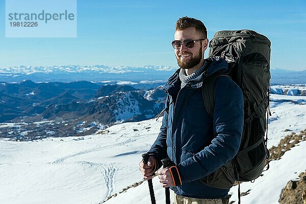 Porträt eines fröhlich lachenden Hipster Reisenden mit Bart und Sonnenbrille in der Natur. Ein Mann beim Wandern in den Bergen mit einem Rucksack und skandinavischen Wanderstöcken im Hintergrund einer Berglandschaft. Reisen Lebensstil Abenteuer Outdoor Erholung