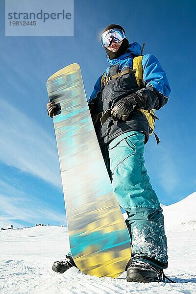 Ein professioneller Snowboarder steht mit seinem Snowboard vor dem blaün Himmel. Weitwinkel. Ansicht von unten