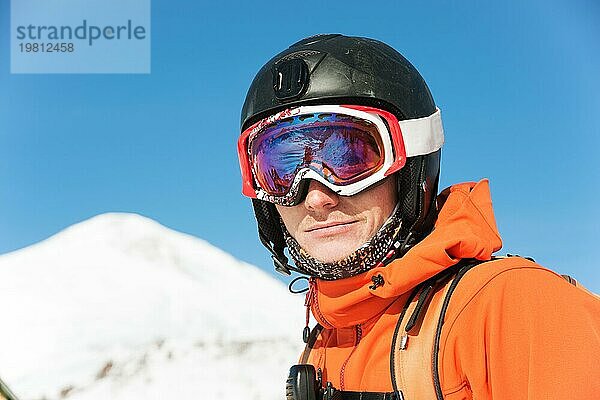 Porträt eines Skifahrers in einem orangefarbenen Overall mit einem Rucksack auf dem Rücken in einem Helm steht vor dem Hintergrund einer schönen kaukasischen Berglandschaft mit schneebedeckten Bergen des kaukasischen Kammes und blaün Himmel. Kopieren Sie das Konzept der aktiven recreati