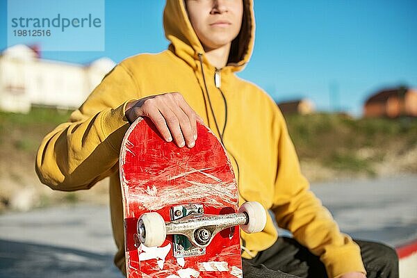 Nahaufnahme eines jungen Hipsters in Sweatshirt und Jeans  der traurig in einem Skatepark sitzt und ein Skateboard hält