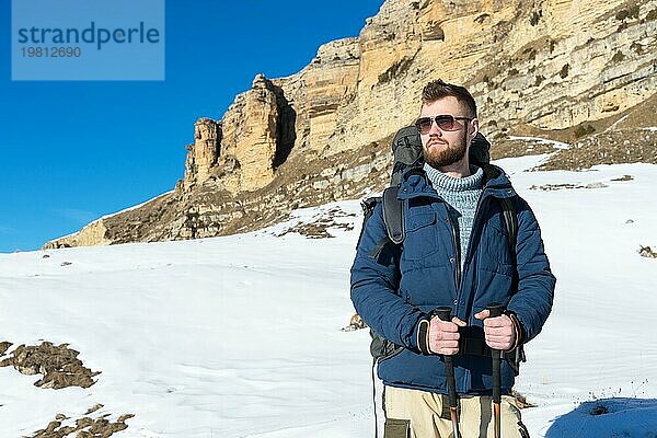 Ein Hipster Reisender mit Bart und Sonnenbrille in der Natur. Ein Mann wandert in den Bergen mit einem Rucksack und skandinavischen Wanderstöcken vor dem Hintergrund einer Berglandschaft und blauem Himmel. Reisen Lifestyle Abenteuer Outdoor Erholung Outdoor Sport