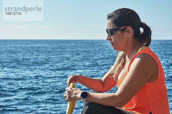 Lateinamerikanische Frau  mittleres Alter  ruht sich aus  stärkt sich  isst  trinkt Wasser  nach einem Fitnessstudio  verbrennt Kalorien  hält sich fit  draußen am Meer  trägt Kopfhörer und Smartwatch