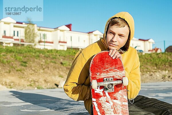 Nahaufnahme eines jungen Hipsters in Sweatshirt und Jeans  der traurig in einem Skatepark sitzt und ein Skateboard hält