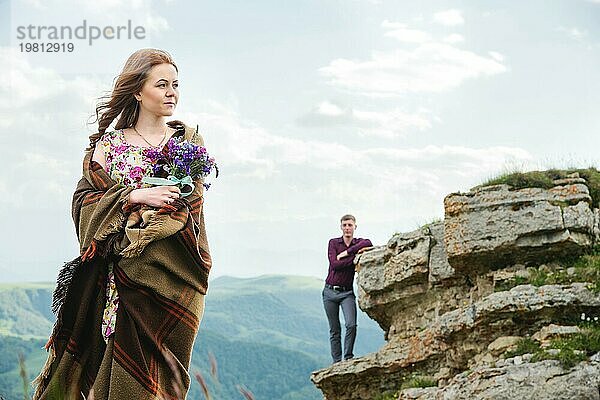 Porträt einer jungen Braut in einem bunten Kleid  eingewickelt in eine Decke und mit einem Strauß Wildblumen in der Natur  die auf dem Hintergrund ihres Freundes steht