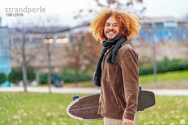 Porträt eines alternativen Mannes mit Skateboard  der in die Kamera lächelt und in einem Park steht