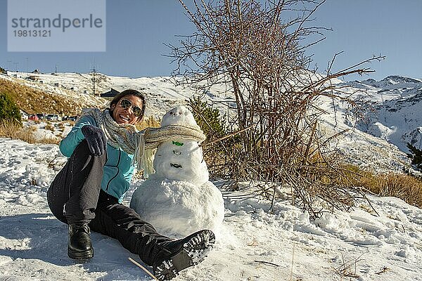 Lateinische Frau sitzt auf gefrorenem Boden neben einem Schneemann