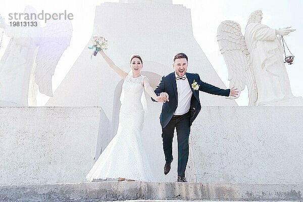 Porträt eines schönen Paares von Jungvermählten an einem Hochzeitstag mit einem Blumenstrauß in den Händen laufen die Treppe hinunter lachend und lächelnd vor dem Hintergrund eines orthodoxen christlichen Denkmal mit Engeln. Das Konzept der christlichen Hochzeit in der Kirche des