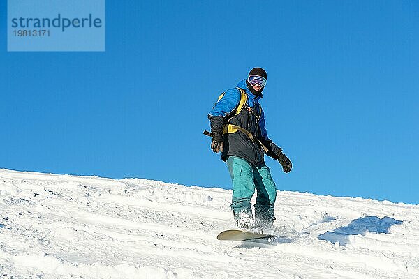 Ein Freeboard Snowboarder mit Skimaske und Rucksack fährt über die schneebedeckte Piste und hinterlässt einen Pulverschnee vor dem blaün Himmel