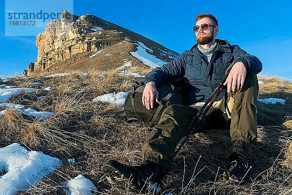 Porträt eines ruhigen Hipster Reisenden mit Bart und Sonnenbrille  der in der Natur sitzt. Ein Mann wandert in den Bergen mit einem Rucksack und skandinavischen Wanderstöcken im Hintergrund eines großen epischen Felsens. Reisen Lifestyle Abenteuer im Freien Erholung im Freien