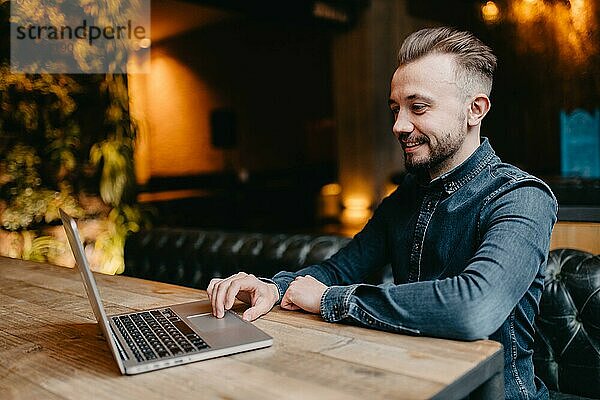 Junger erfolgreicher lächelnder Freiberufler im Jeanshemd  der an einem Laptop in einer Kokerei arbeitet