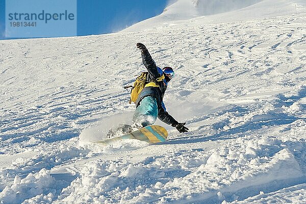 Ein Freeboard Snowboarder mit Skimaske und Rucksack fährt über die schneebedeckte Piste und hinterlässt einen Pulverschnee vor dem blaün Himmel