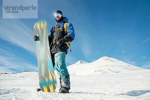 Ein professioneller Snowboarder steht mit seinem Snowboard vor der Kulisse des schlafenden Vulkans Elbrus. Nordkaukasus. Beschaffung für Werbung oder Plakat mit Copy Space. Konzept von Wintersport und Erholung