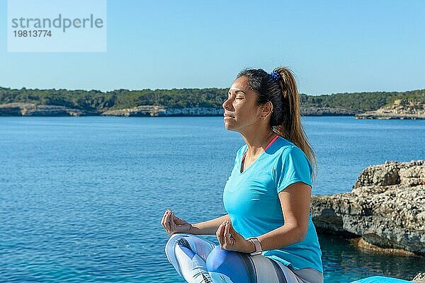 Frau in einer sitzenden Yogahaltung beim Meditieren am Meer an einem sonnigen Tag