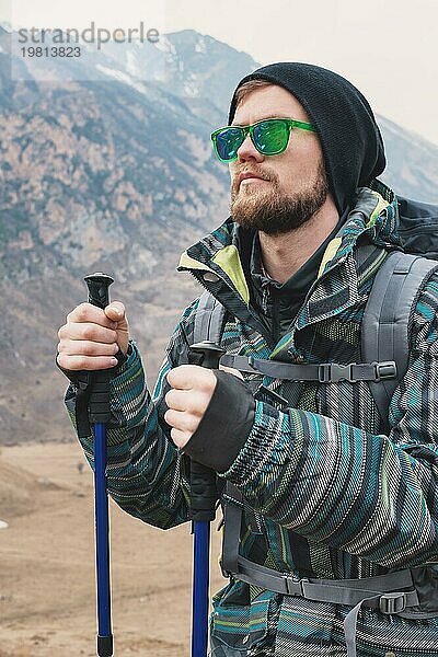 Ein Mann mit Bart und Sonnenbrille in einer Membranjacke  Hut  mit einem Rucksack und Stöcken für Nordic Walking  ein Reisender  der in der freien Natur steht und auf die Berge schaut