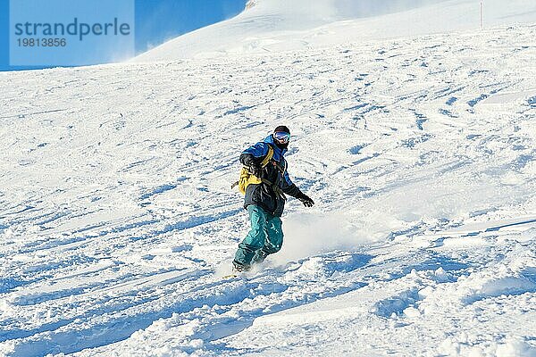 Ein Freeboard Snowboarder mit Skimaske und Rucksack fährt über die schneebedeckte Piste und hinterlässt einen Pulverschnee vor dem blaün Himmel