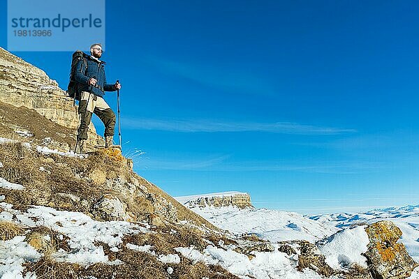 Ein Hipster Reisender mit Bart und Sonnenbrille in der Natur. Ein Mann wandert in den Bergen mit einem Rucksack und skandinavischen Wanderstöcken vor dem Hintergrund einer Berglandschaft und blauem Himmel. Reisen Lifestyle Abenteuer Outdoor Erholung Outdoor Sport