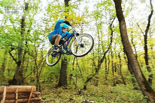 Ein junger Fahrer mit Helm und blauem Sweatshirt fliegt auf einem Fahrrad  nachdem er von einem hohen Kicker auf einem Waldradweg gesprungen ist. Aufnahme mit Langzeitbelichtung Verdrahtung