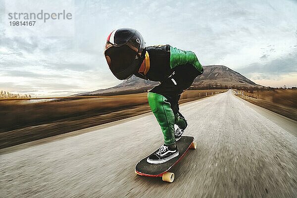 Ein junger Mann in einem Fullface Helm und einem Lederanzug in einem speziellen Gestell fährt ein Longboard auf afsaltu Sonnenuntergang im Hintergrund Berge und schönen Himmel mit hoher Geschwindigkeit