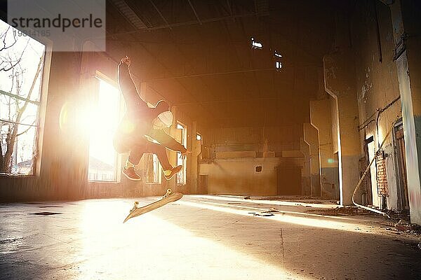 Ein junger Skater mit weißer Mütze und schwarzem Sweatshirt macht einen Trick mit einem Skatesprung in einem verlassenen Gebäude im Gegenlicht der untergehenden Sonne. Das Konzept der jugendlichen Untergrundkultur und des Skateboarding