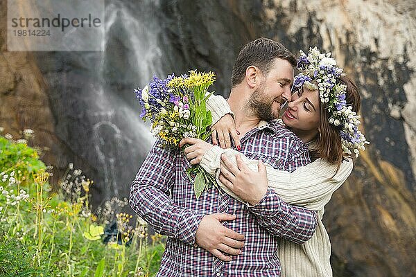 Ein junges Paar von verliebten Hipstern spaziert in der Natur vor dem Hintergrund eines Wasserfalls und Bergkräutern und Blumen