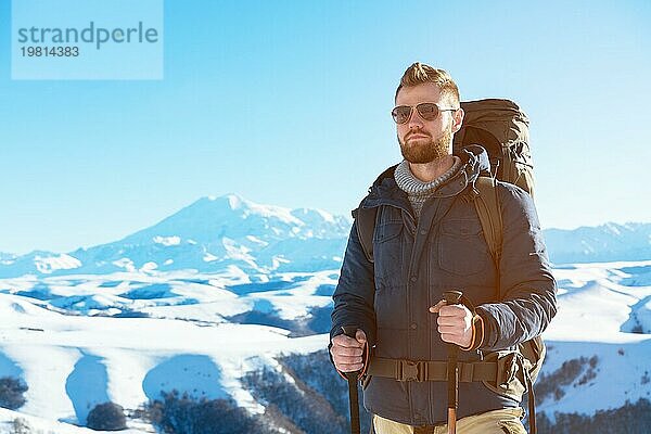 Ein Hipster Reisender mit Bart und Sonnenbrille in der Natur. Ein Mann wandern in den Bergen mit einem Rucksack und skandinavischen Wanderstöcke im Hintergrund eines Berges Elbrus Landschaft und blaün Himmel. Reisen Lifestyle Abenteuer Outdoor Erholung übertreffen