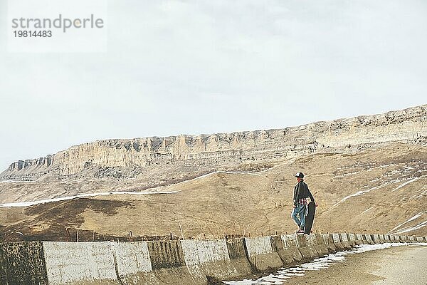 Stylische rothaarige Frauen in Jeansoveralls stehen mit einem Longboard auf einer Bodenwelle am Highway in den Bergen