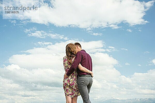 Blick von hinten. Junges Paar umarmt Küsse gegen den blaün Himmel und weißen Wolken. Das Konzept der idealen Beziehungen und Familie
