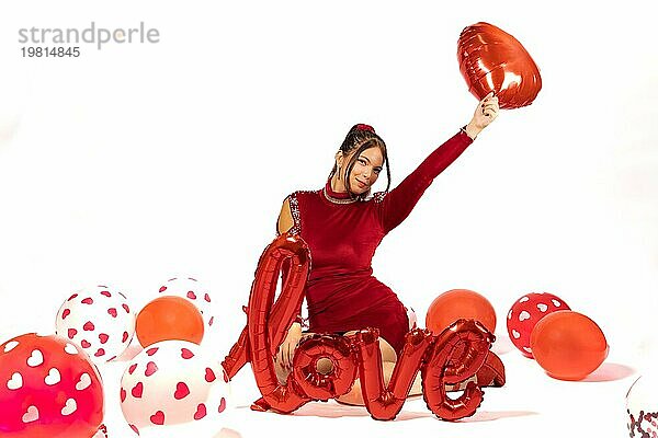Schöne elegante Mädchen im roten Kleid von Luftballons auf weißem Hintergrund mit Liebe Zeichen umgeben. Valentinstag Konzept. Rote und weiße Farbpalette