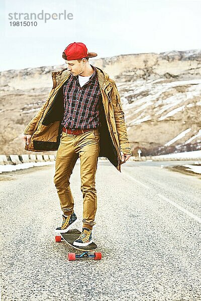 Stylish happy Junger Mann in Mütze und Hose Jogger rollen auf einem Longboard eine Bergstraße hinunter  genießen das Leben