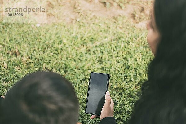 Schöne lateinische Familie lachend von Smartphone Videos zu beobachten  sitzen auf der grünen Park zwei Brüder und die Mutter an einem sonnigen Tag
