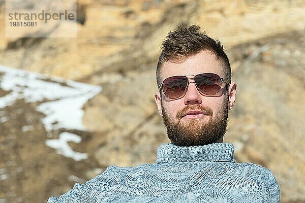 Porträt eines Reisenden Hipster mit Sonnenbrille und einem gestrickten grauen Pullover ruht vor dem Hintergrund eines Winters Felsen  Das Konzept der Entspannung in den Bergen und Trekking mit Wandertouren