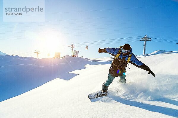 Ein Snowboarder mit Skimaske und Rucksack fährt auf einer schneebedeckten Piste und hinterlässt einen Pulverschnee vor dem blaün Himmel und der untergehenden Sonne. Foto in Bewegung