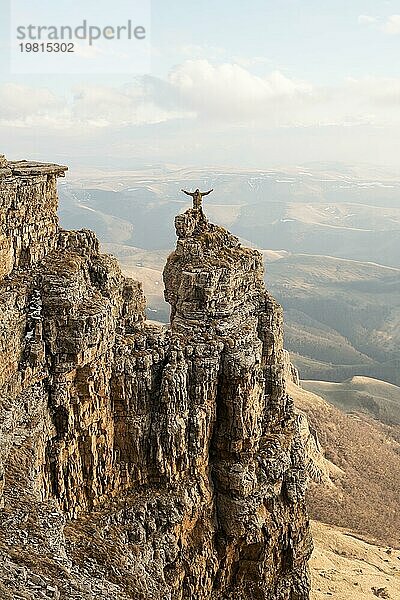 Ein glücklicher Mann mit hoch erhobenen Händen steht auf einem separat stehenden Felsen  der sich über den Wolken befindet  vor dem Hintergrund von Tälern  Hügeln  Sonnenuntergang und einem schönen Himmel